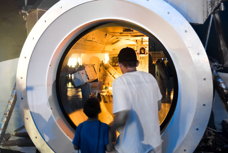 Kai and Kenny looking into the inside of the Lunar Excursion Module (LEM), looking into the very small space where two humans trusted their lives utterly to the machinery. Trusted their lives to metal, fabric, electricity, plastic, and propellant. Came down uncannily from out of a black daytime sky to alight softly upon the dusty surface of another world.